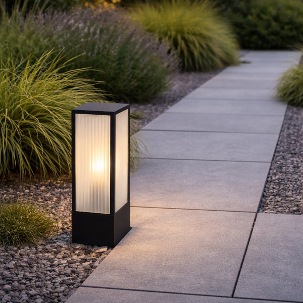 Moderne schwarze Sockelleuchte mit warmem Licht an einem Gartenweg aus grauen Steinplatten und Ziergräsern.