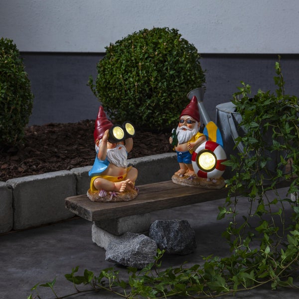 Zwei Gartenzwerge mit Sonnenlicht sitzen auf einer Holzbank in einem Garten.