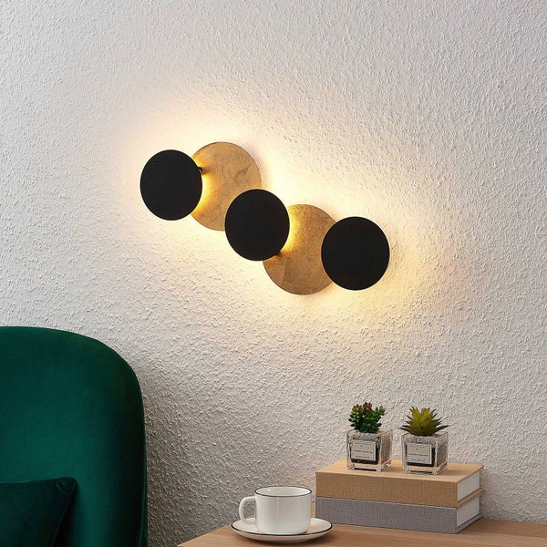 Moderne Wandleuchte mit schwarzen und goldfarbenen Scheiben. Indirektes Licht über einem Tisch mit Pflanzen und Dekoration.