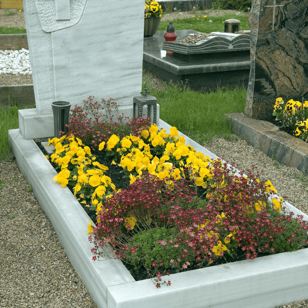 Gepflegtes Grab mit weißem Marmorgrabstein und Einfassung, bepflanzt mit gelben Stiefmütterchen und roten Blumen.