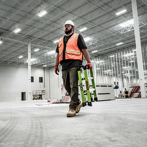 Handwerker mit Schutzkleidung und Schutzhelm trägt eine zusammenklappbare Stehleiter durch eine große, offene Baustelle mit Metallständerwänden.
