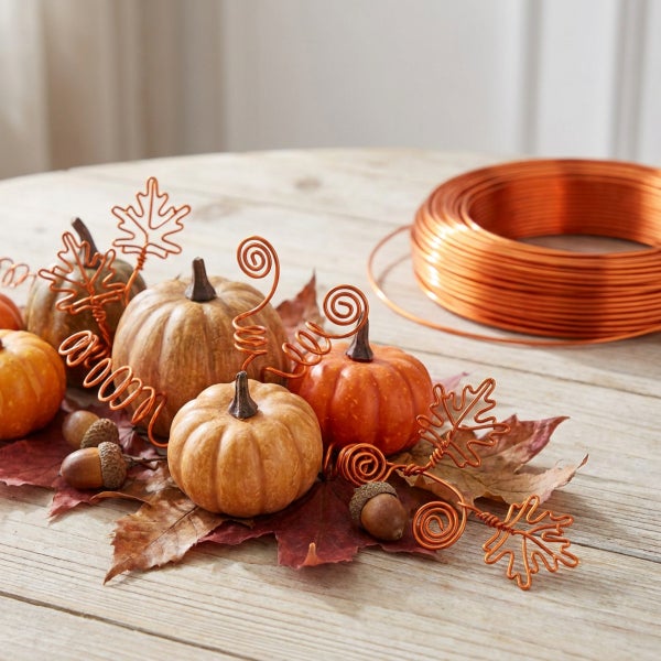 Herbstliche Dekoration mit künstlichen Kürbissen, Eicheln, Herbstblättern und einer Rolle Kupferdraht auf einem Holztisch.