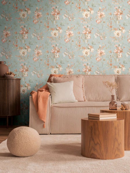 Wohnzimmer mit Vliestapete mit Blumenmuster, beigem Cordsofa, Holztischen und einer runden Sitzgelegenheit vor einer gemusterten Wand.