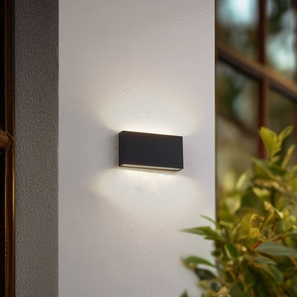 Schwarze rechteckige LED-Außenwandleuchte mit Up-and-Down-Lichteffekt montiert an einer Hausfassade neben Pflanzen.