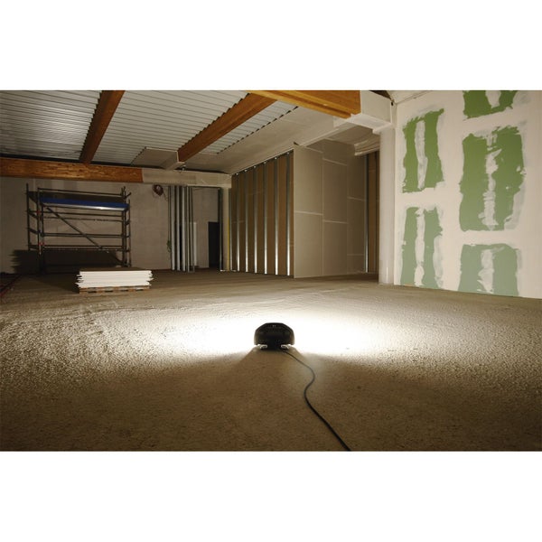 Ein Baustrahler beleuchtet den Boden in einem Raum im Rohbau mit Trockenbauwänden und einem Gerüst im Hintergrund.