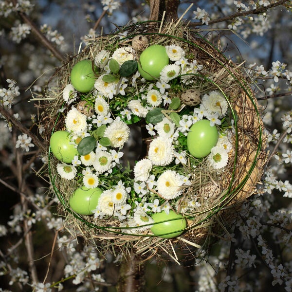 Osterkranz aus Stroh dekoriert mit Ostereiern und Frühlingsblumen hängend an einem blühenden Baum im Freien