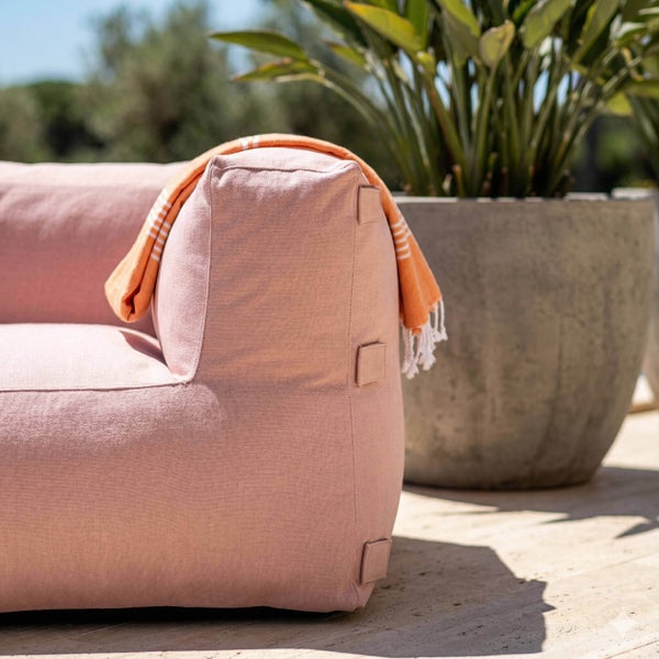 Rosafarbenes Outdoor-Loungemöbel aus strukturiertem Stoff mit orangefarbener Decke auf einer sonnigen Terrasse neben einer Pflanze im Topf.