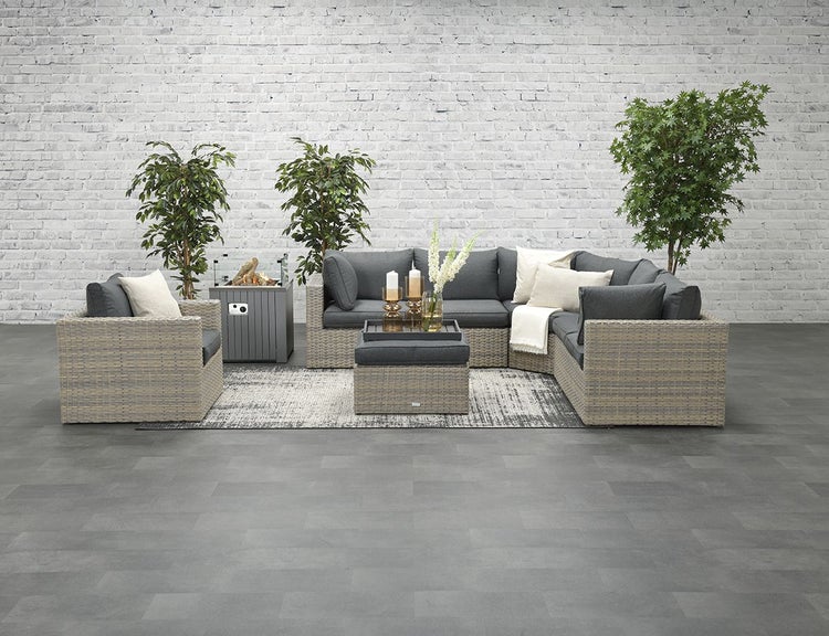 Modernes Garten-Lounge-Set aus Polyrattan mit Ecksofa, Sessel, Hocker und Feuertisch auf einem Teppich vor einer Ziegelwand.
