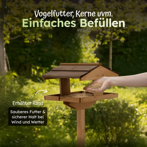 Ein hölzernes Vogelfutterhaus auf einem Pfosten mit erhöhtem Rand und einer Hand, die Körner in die Futterschale streut.