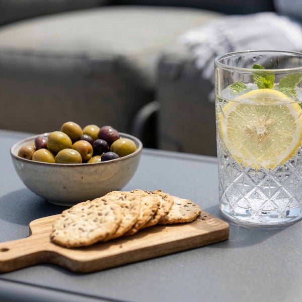 Oliven in einer Schale, Cracker auf einem Holzbrett und ein Glas Wasser mit Zitrone auf einem hellen Gartentisch im Außenbereich.