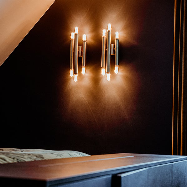 Zwei Wandleuchten mit mehreren vertikalen Lichtstäben beleuchten eine dunkle Wand in einem Schlafzimmer.
