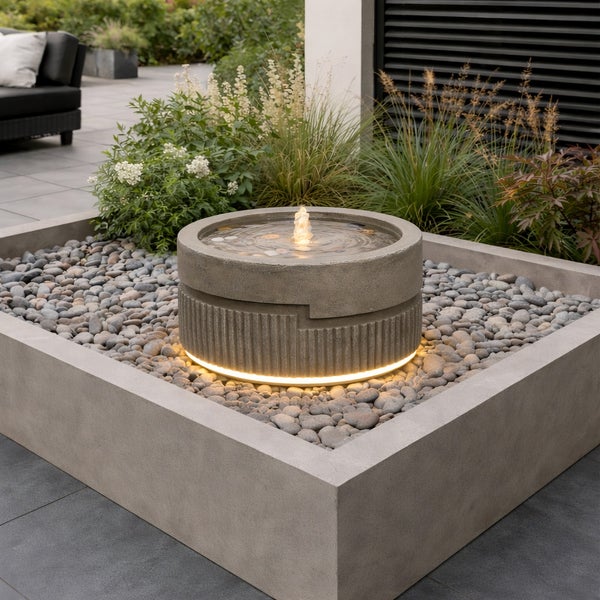 Moderner runder Gartenbrunnen in Steinoptik mit LED-Beleuchtung, in einem Kiesbett aus Kieselsteinen und einer quadratischen Betoneinfassung.