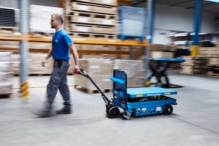 Ein Mann zieht einen fahrbaren elektrischen Scherenhubwagen von HanseLifter durch ein organisiertes Lager mit Hochregalen.