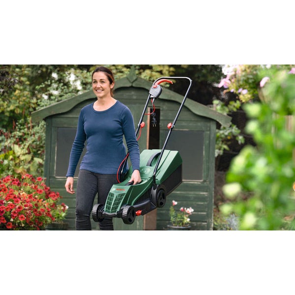 Eine Frau trägt einen grünen Bosch Elektro-Rasenmäher in einem Garten mit Blumen und Gartenhaus.