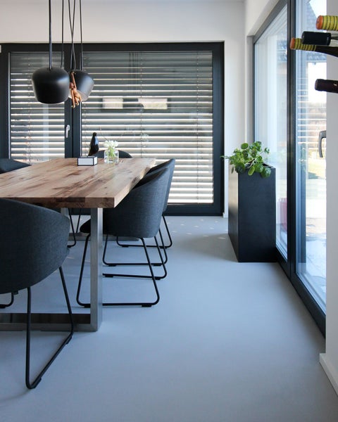 Modernes Esszimmer mit massivem Holztisch, dunkelgrauen Polsterstühlen und großen Fenstern mit Jalousien auf fugenlosem grauem Boden.