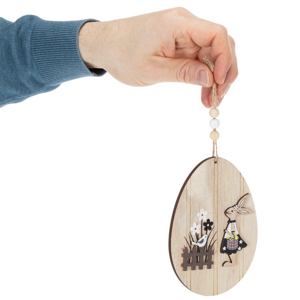 Hand hält österlichen Eieranhänger aus Holz mit Hasenmotiv, Blumen und Zaun.