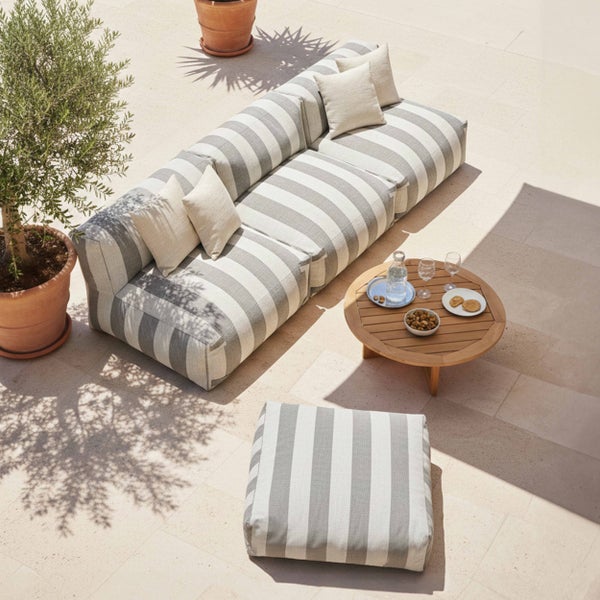 Gartenlounge-Set mit grau-weiß gestreiftem Modulsofa, passendem Hocker, rundem Holztisch und Olivenbaum auf einer sonnigen Steinterrasse.