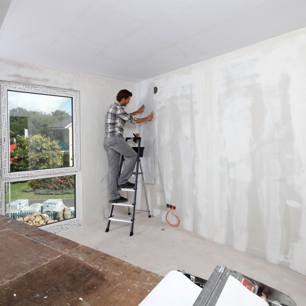 Mann auf einer Leiter, der eine Wand tapeziert in einem hellen Raum.
