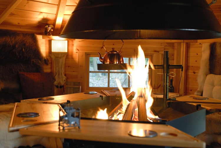 Helle Holzhütte mit offenem Kamin und Grillstelle in der Mitte, dekoriert mit Fellen und einer Teekanne über dem Feuer.