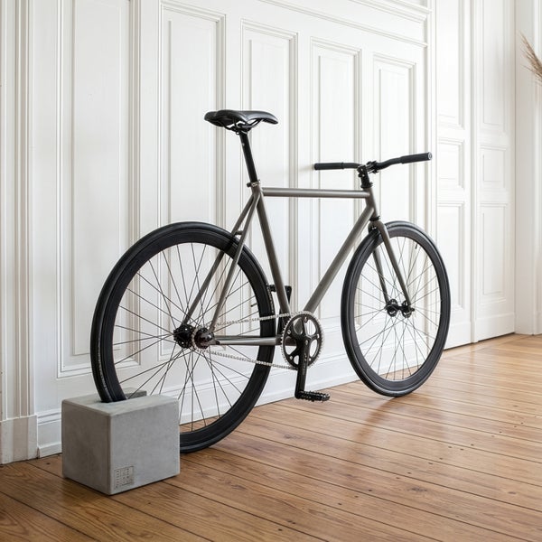 Minimalistischer Fahrradständer aus Beton in Würfelform hält ein graues Fahrrad am Hinterrad in einem hellen Raum mit Holzboden.