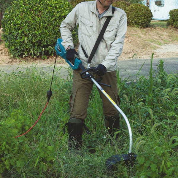 Eine Person benutzt einen elektrischen Rasentrimmer mit Schultergurt zum Schneiden von hohem Gras im Garten.