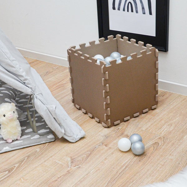 Quadratische Box aus Schaumstoff-Puzzlematten gefüllt mit weißen und silbernen Bällen, auf hellem Holzboden neben einem Spielzelt.