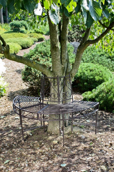 Halbrunde Baumbank aus Metall, um einen Baumstamm in einer Gartenanlage platziert.