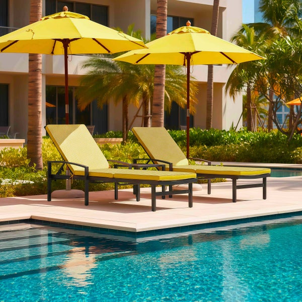 Sonnige Poollandschaft mit zwei dunklen Gartenliegen, gelben Auflagen und zwei großen gelben Sonnenschirmen auf einer hellen Steinterrasse.