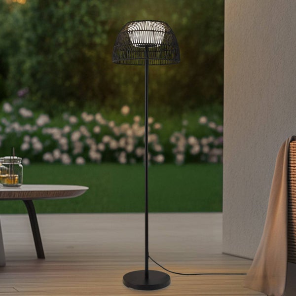 Moderne Außenstehleuchte mit schwarzem Gestell und geflochtenem Schirm auf einer Holzterrasse vor einem Garten bei Dämmerung.