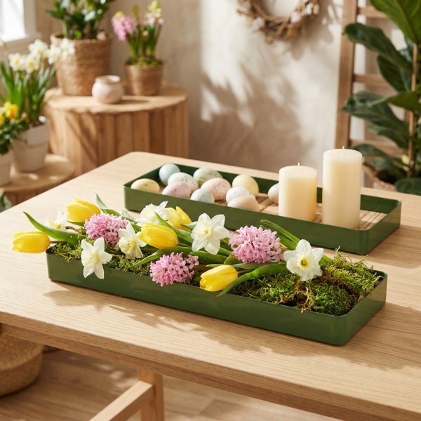 Zwei längliche grüne Deko Tabletts aus Metall auf einem Holztisch, arrangiert mit Frühlingsblumen, Moos, Kerzen und Ostereiern als Tischschmuck.