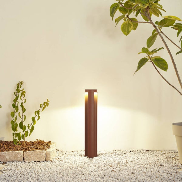 Moderne zylindrische Gartenleuchte mit warmweißem Licht auf hellem Kiesboden zwischen Pflanzen.