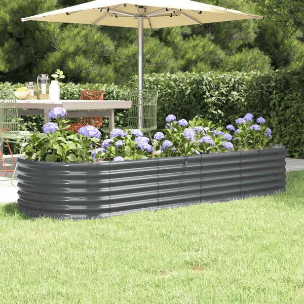 Ovales Hochbeet aus anthrazitfarbenem Wellblech auf einer Rasenfläche, bepflanzt mit violetten Blumen, Gartenmöbel im Hintergrund.