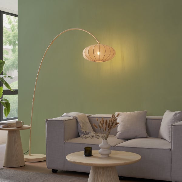 Moderne Bogenstehleuchte mit geripptem Stoffschirm in einem hellen Wohnzimmer mit grauem Sofa und runden Holzbeistelltischen.