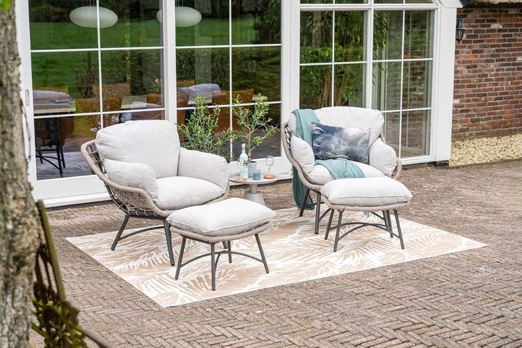 Zwei Garten-Loungesessel mit Fußhockern und Polsterauflagen auf einem gemusterten Outdoor-Teppich auf einer hellen, gepflasterten Terrasse.
