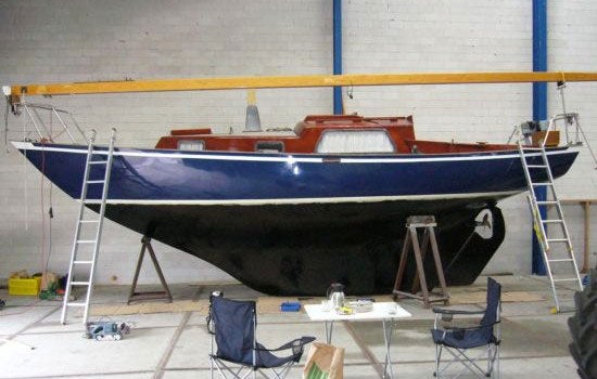 Ein Segelboot auf einem Bock in einer Werkstatt mit Leitern an beiden Seiten und einem Tisch mit zwei Klappstühlen im Vordergrund.