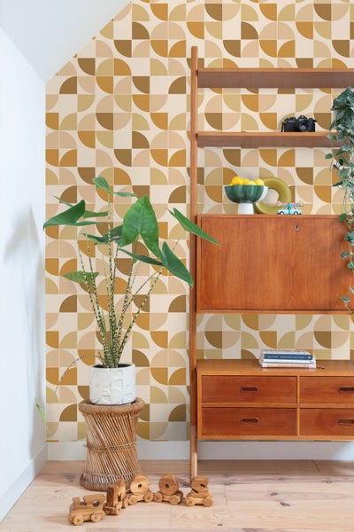 Vliestapete mit geometrischem Muster aus ockerfarbenen und beigen Halbkreisen in einem hellen Raum mit Holzmöbeln und Zimmerpflanze.