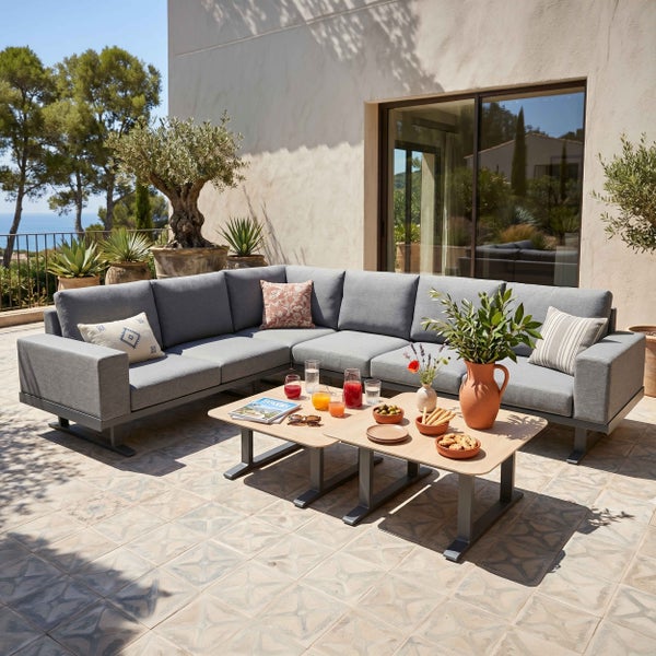 Moderne L-förmige Garten-Lounge-Ecke mit Polstern und zwei Kaffeetischen auf einer sonnigen Terrasse mit mediterranen Pflanzen.