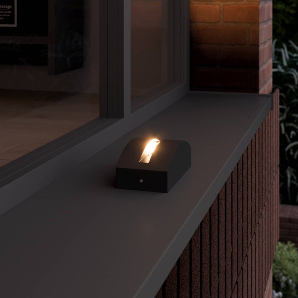 Rechteckige schwarze Außenleuchte auf einer Fensterbank an einer Ziegelwand, warmes Licht strahlt aus einer schmalen Öffnung nach oben.