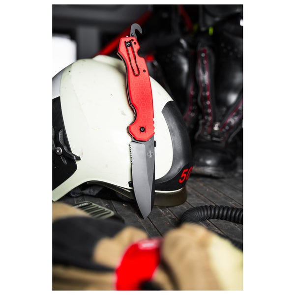 Rettungsmesser mit Wellenschliff, Glasbrecher und Gurtschneider neben einem Feuerwehrhelm und Stiefeln.