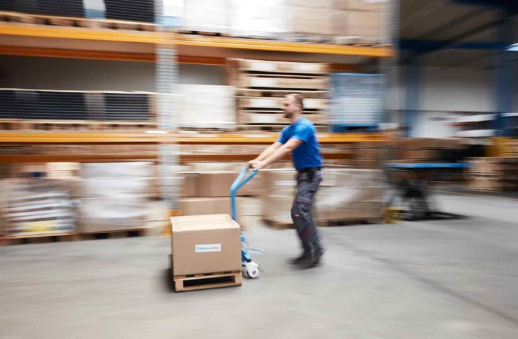 Ein Mann transportiert einen Karton auf einer Palette mit einem blauen Hubwagen in einem Lager mit Regalsystemen.