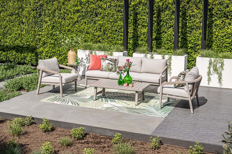 Modernes Garten-Lounge-Set aus Sofa, Sesseln und Couchtisch auf einer hellen, organisierten Terrasse mit Outdoorteppich und Hecke.