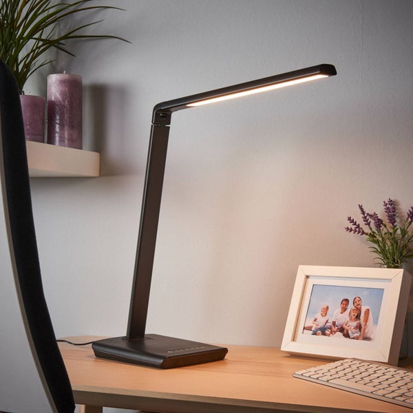 Moderne schwarze LED-Schreibtischleuchte auf einem Holzschreibtisch neben einem Bilderrahmen und einer Tastatur.