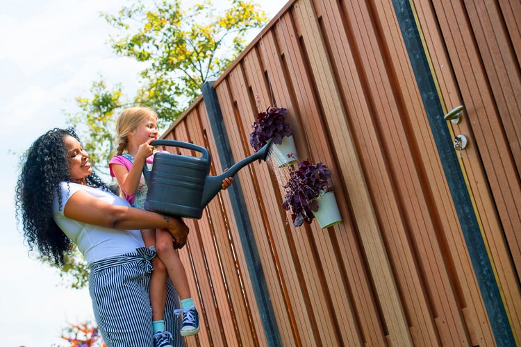 Eine Frau hält ein Kind, das mit einer Gießkanne Pflanzen vor einem Holzzaun gießt.