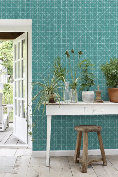Raumansicht mit geometrischer Tapete, weißem Tisch mit Pflanzen und Holzschemel