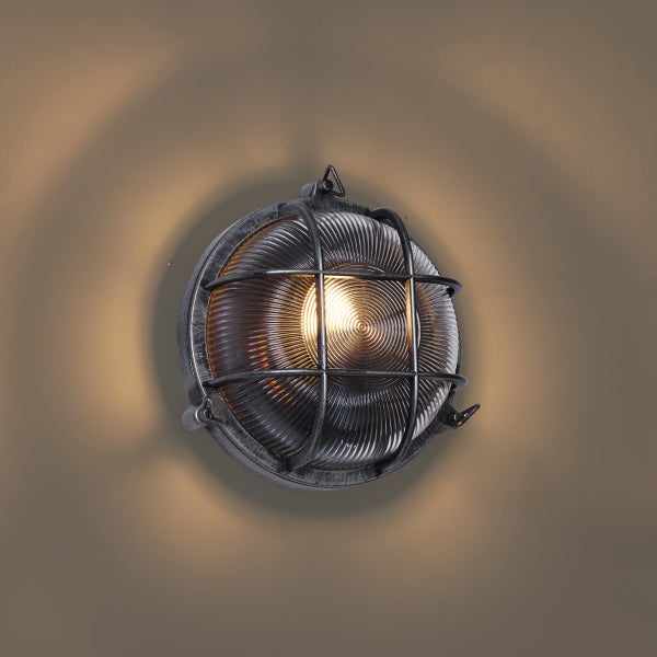 Runde Wandleuchte im industriellen Stil aus grauem Metall mit Schutzgitter und geripptem Glas, beleuchtet.