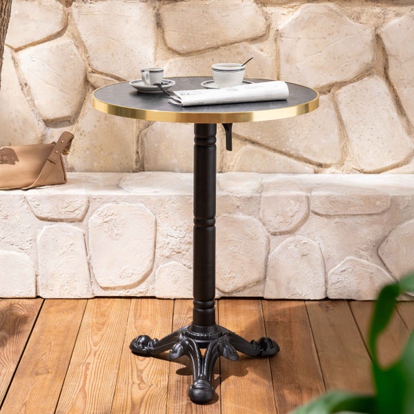 Runder Bistrotisch mit schwarzem Metallfuß und goldfarbenem Rand auf einer Terrasse vor einer Steinmauer.