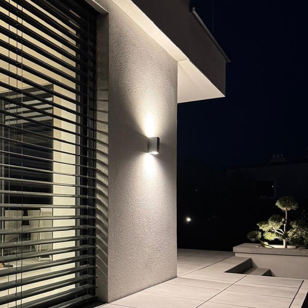 Moderne rechteckige Außenwandleuchte an einer verputzten Fassade bei Nacht mit Up-and-Down-Lichteffekt neben einem Fenster mit Jalousien.