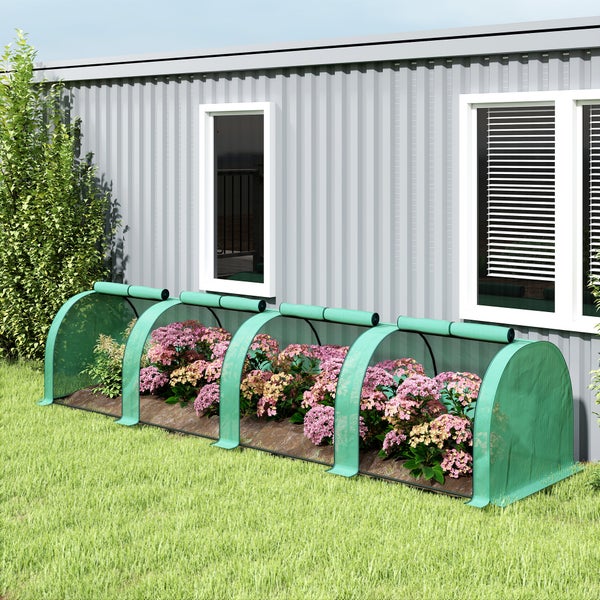 Grüner Pflanzschutztunnel aus Netzgewebe über blühenden Blumen im hellen Garten vor einem Bambuszaun und einer Mauer.