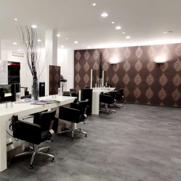 Moderner Friseursalon mit dunklem Boden, weißen Arbeitstischen und einer braunen gemusterten Tapete an der Akzentwand.