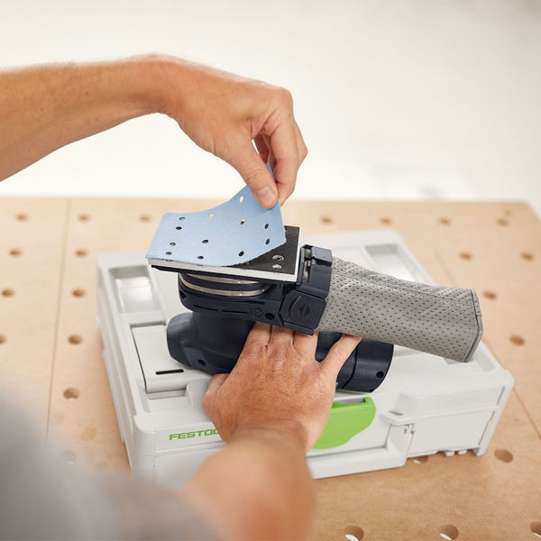 Eine Person befestigt Schleifpapier an einem Schwingschleifer von Festool, der auf einem Werkzeugkoffer derselben Marke steht.